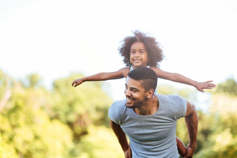 A father carries a young girl on his back. She has her arms spread wide. They are outdoors but the background is blurred.
