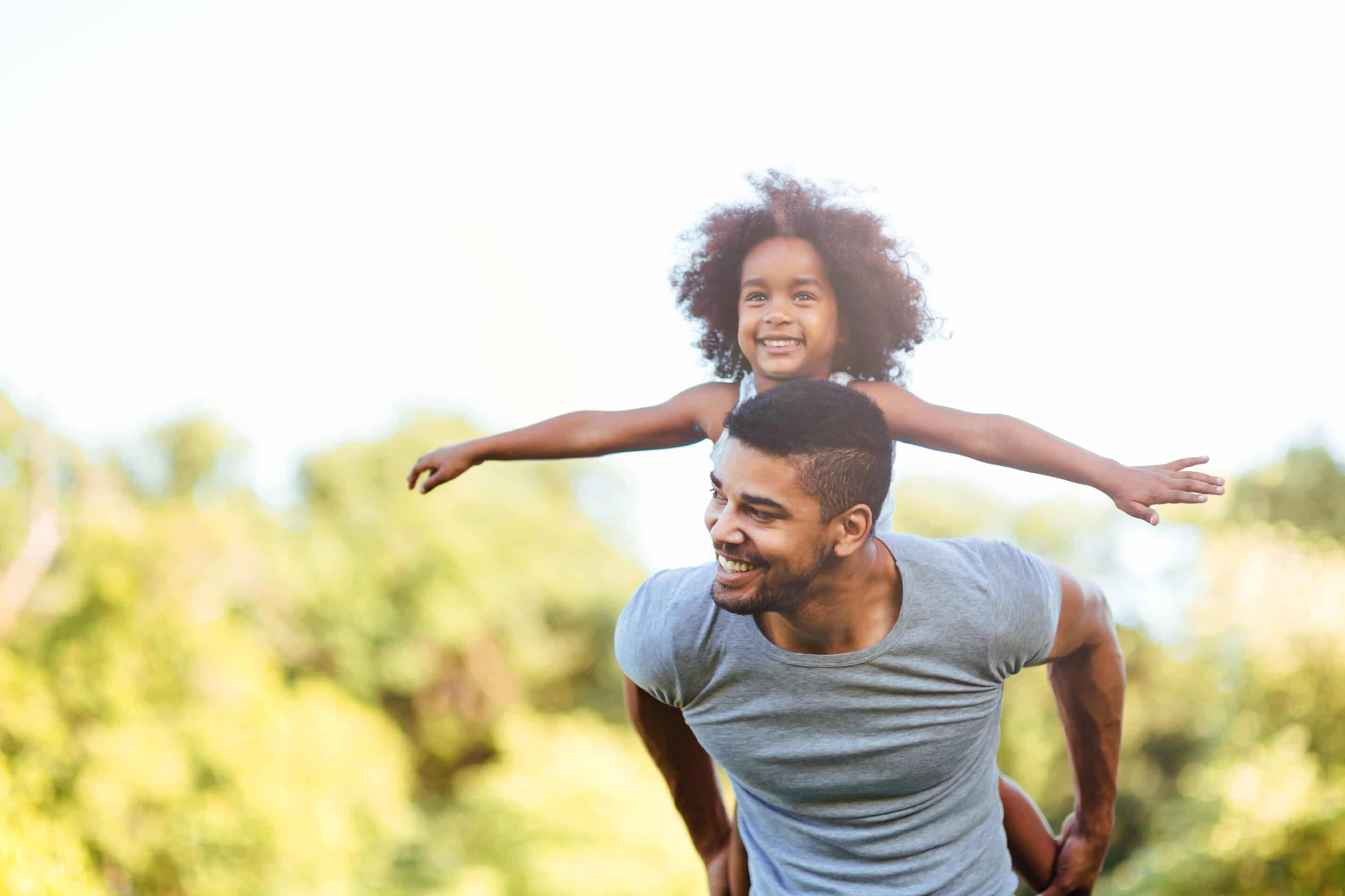 A father carries a young girl on his back. She has her arms spread wide. They are outdoors but the background is blurred.