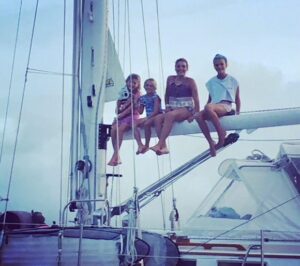 Four people sitting on a sailboat's boom against a cloudy sky.