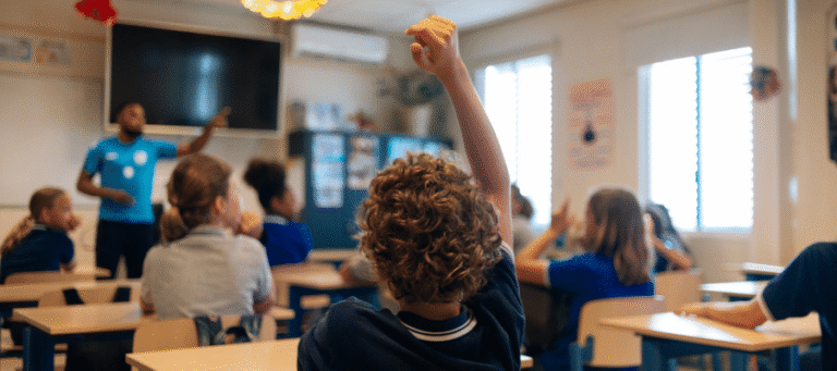 A student raising hand in a classroom full of children.