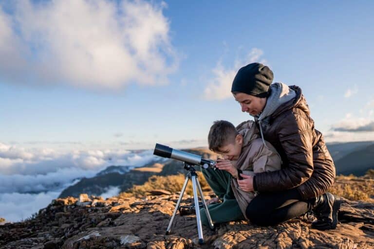 A woman and child using a telescope on a rocky mountain under a blue sky.