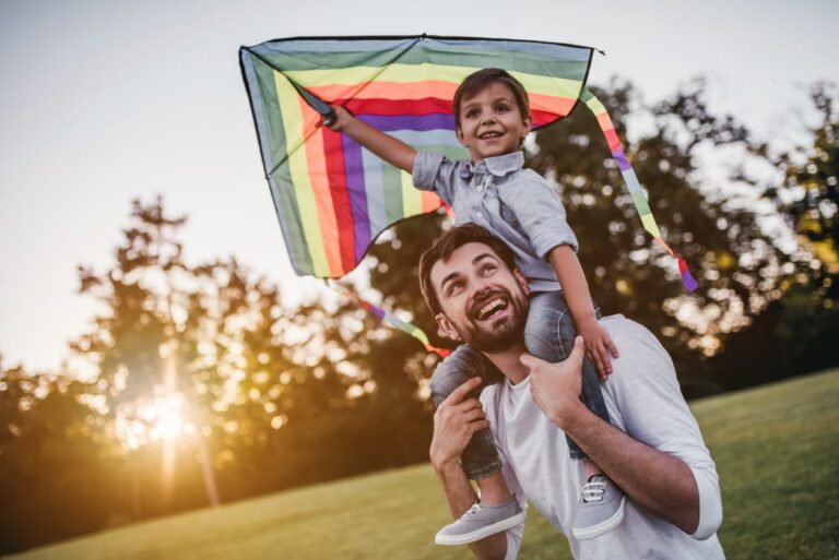 Father carrying son on shoulders flying a colorful kite at sunset.