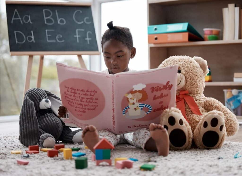 A young girl reads a large storybook in a cozy room surrounded by toys.